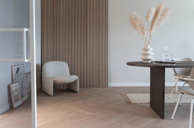 Luxe visgraat pvc-vloer in een minimalistisch interieur met donkere houten wandpanelen, designfauteuil, ronde eetkamertafel en decoratieve vaas met pampasgras.