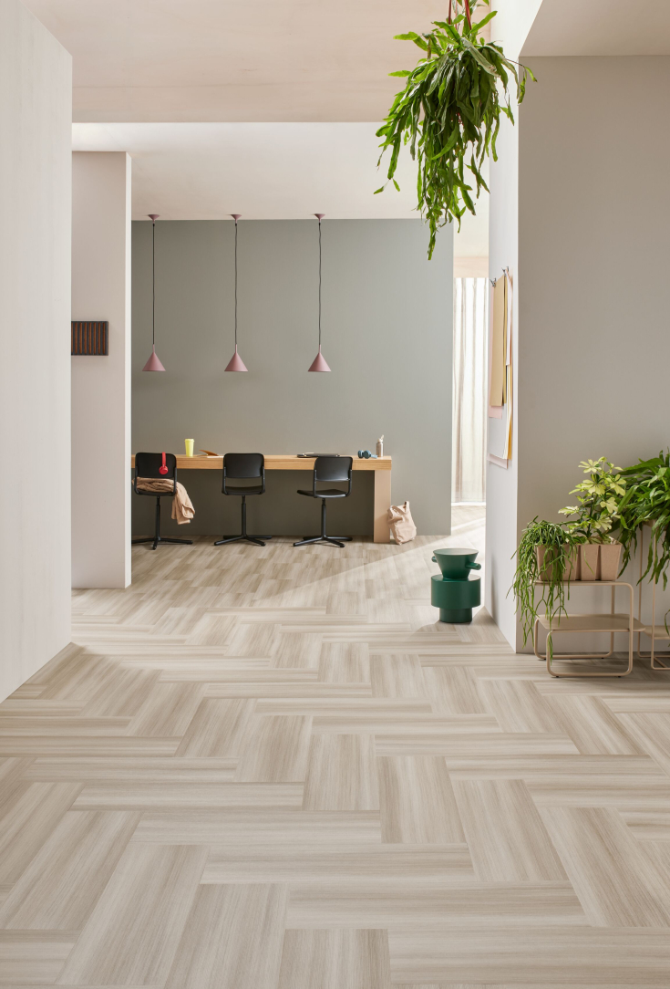 Lichte plak pvc-tegelvloer in visgraatpatroon binnen een modern en groen kantoorinterieur met een houten bureautafel, zwarte bureaustoelen, hangplanten en zachte pastelaccenten.