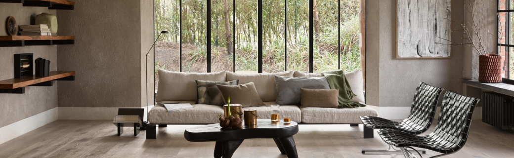 Lichte eiken klik pvc-vloer in een sfeervolle woonkamer met groot raam, modern designmeubilair, zwart houten salontafel en een comfortabele beige bank met decoratieve kussens.