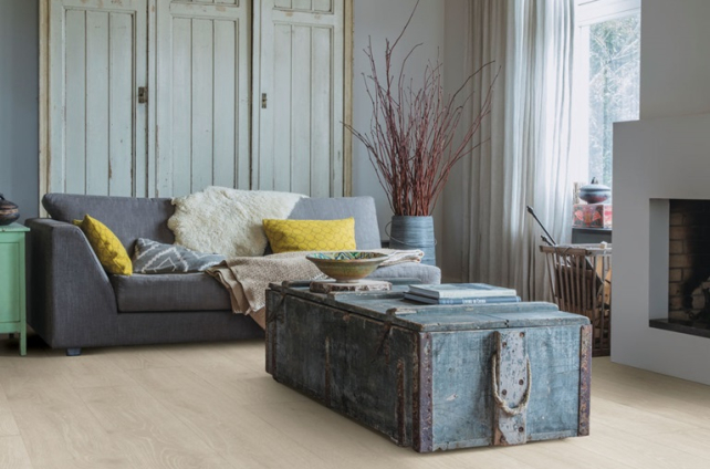 Sfeervolle woonkamer met lichte houtlook PVC vloer van Quick-Step en een warme inrichting met vintage kast en grijze bank.