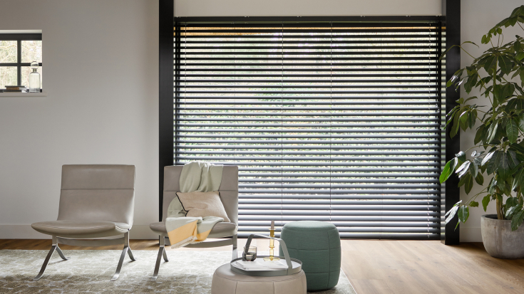 Moderne woonkamer met brede pvc-vloer in warme houttint, zwarte jaloezieën en minimalistisch interieur met designstoelen en een groene poef.