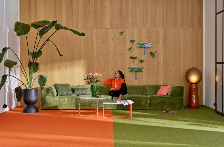 Kleurrijke woonkamer met groen en oranje tapijt, groene fluwelen bank en grote kamerplant voor houten wand