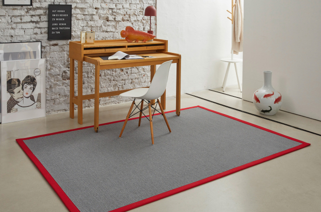 Modern grijs vloerkleed met rode rand onder houten bureau en witte stoel in industriële werkruimte