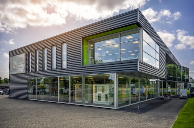 Vooraanzicht van de showroom van Groen Deventer Zonwering met geparkeerde auto's en reclamebord met zonwering